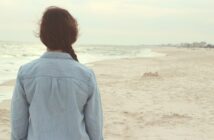 Frau mit Jeanshemd und geflochtenem Zopf von hinten mit Blick vom Strand aus auf das Meer