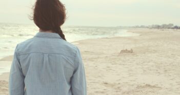 Frau mit Jeanshemd und geflochtenem Zopf von hinten mit Blick vom Strand aus auf das Meer
