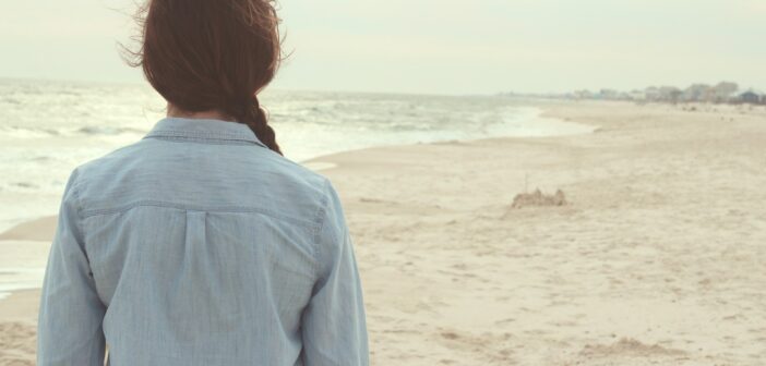 Frau mit Jeanshemd und geflochtenem Zopf von hinten mit Blick vom Strand aus auf das Meer