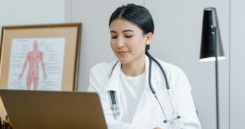 Junge Ärztin sitz vor ihrem Laptop. Um den Hals hängt ihr ein Stethoskop