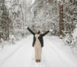 Frau steht auf winterlichem Weg im Wald und reckt die Arme in die Höhe.